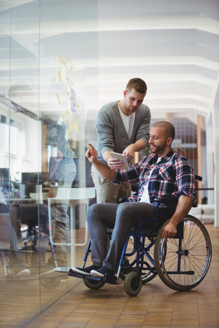 Uomo d'affari con disabilità discute con un collega mentre applica note adesive su una finestra di vetro in un ufficio creativo.